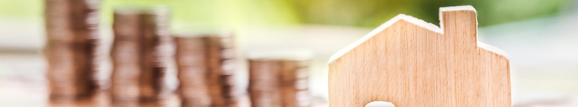 Symbolfoto Steuern und Abgaben: ein Spielzeughaus aus Holz, im Hintergrund, unscharf, gestapelte Münzen.